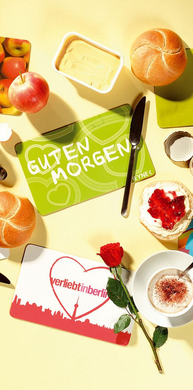 Frühstückstisch mit einem grünen Frühstücksbrettchen, Brötchen, Obst, Joghurt und einer roten Rose auf gelbem Untergrund.